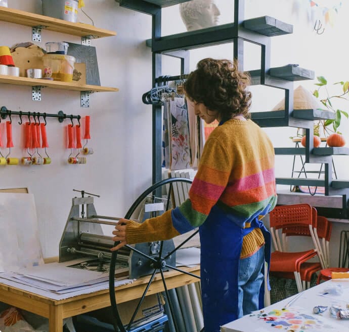 A person wearing a colorful sweater and blue apron operates a printing press in a bright, organized art studio. Shelves with supplies and artwork adorn the walls, and light pours in from a window, highlighting the creative workspace.